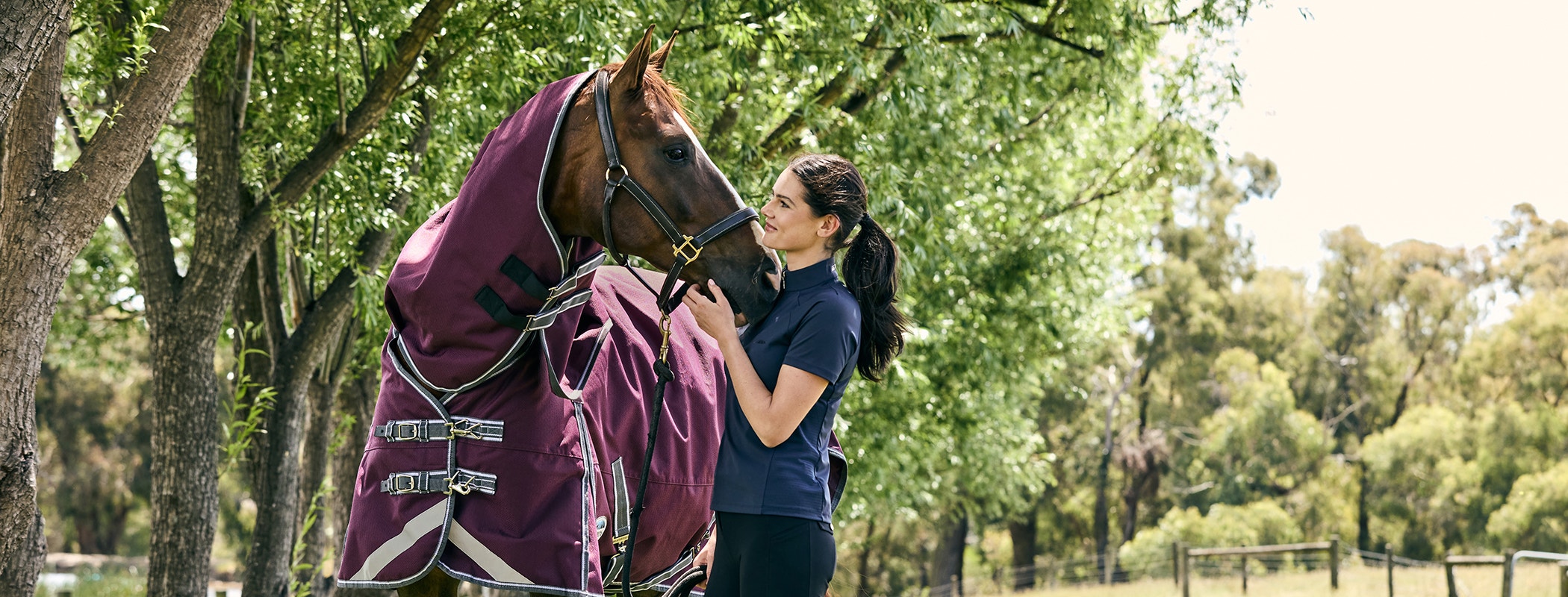 horse rider and horse stood outside under trees. The rider is wearing a short sleeve weatherbeeta top. The horse is wearing a burgundy weatherbeeta turnout rug