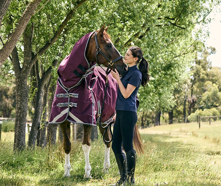 horse rider and horse stood outside under trees. The rider is wearing a short sleeve weatherbeeta top. The horse is wearing a burgundy weatherbeeta turnout rug